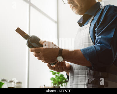 Man holding a red wine bottle and reading the label, he is cooking in the kitchen at home Stock Photo