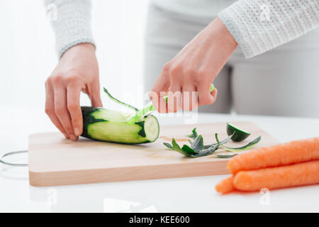 Woman cooking healthy food in her kitchen, she is peeling a fresh cucumber on the chopping board Stock Photo