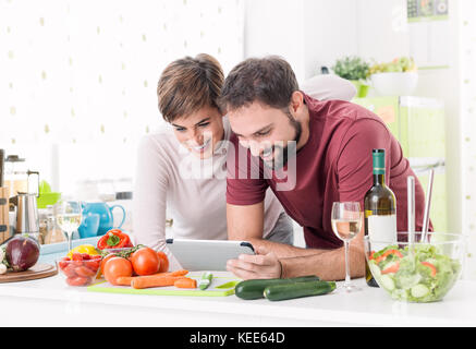Young smiling couple cooking together at home, they are preparing vegetables and checking recipes online using a digital touch screen tablet Stock Photo