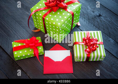 Bunch of colorful presents, wrapped in paper and tied with red ribbon and bows, with an opened envelope and blank paper, on an old wooden table. Stock Photo