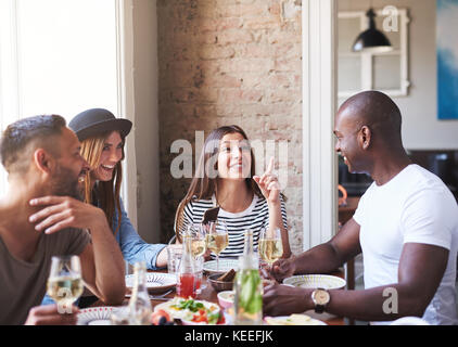 Diverse small group of four happy friends having wine and dinner together in restaurant with brick wall and bright large window in background Stock Photo