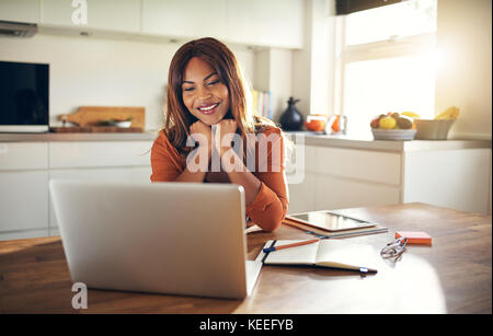 Smiling young African female entrepreneur sitting at her kitchen table using a laptop while working from home Stock Photo