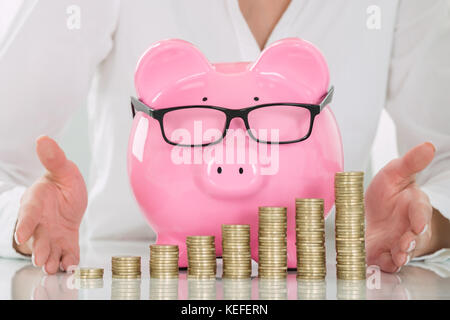 Close-up Of A Female's Hand Protecting Piggybank And Stacked Coins On Desk Stock Photo