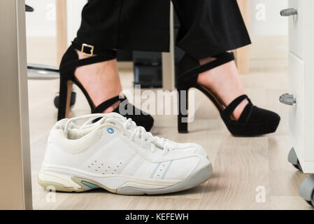 Pair Of A White Sport Shoes Besides Businesswoman's Foot Wearing High Heels Stock Photo