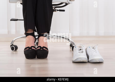 Pair Of A White Sport Shoes Besides Businesswoman's Foot Wearing High Heels Stock Photo