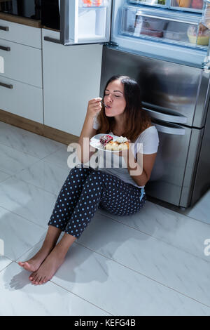 Young Woman Enjoy Eating Sweet Food While Sitting On Floor In Kitchen Stock Photo