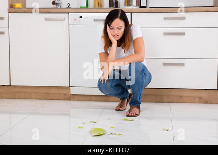 Young Unhappy Woman Looking At The Broken Plate Fallen On Floor In The Kitchen Stock Photo