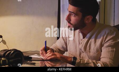 Stylish young man sitting at his desk at home at night, working on paperwork taking notes and making amendments in front of computer Stock Photo
