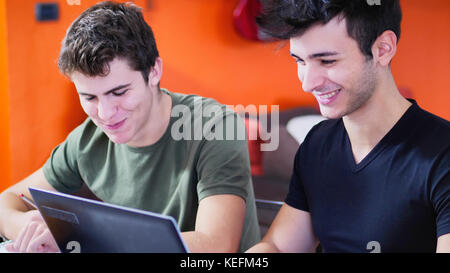 Two young friends doing homework with laptop computr and tablet PC while sitting at table with notebooks. Stock Photo