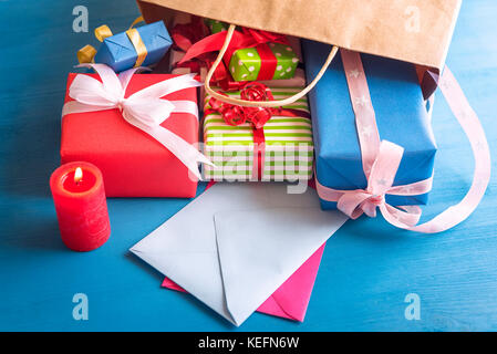 Many and colorful presents spilled out of a bag on a blue wooden table, falling over empty envelopes, near a lit red candle. Stock Photo