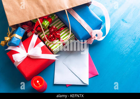 Brown paper bag overturned on a blue wooden table, while multicolored presents and closed envelopes come out of it, near a red, lit candle. Stock Photo