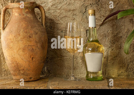 an old clay jug and a bottle of white wine with a wineglass stand on the background of a clay wall Stock Photo