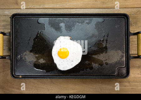 Fried egg white and yolk rectangular cast iron flat griddle pan with wooden handles, wood tabletop background, flat lay Stock Photo