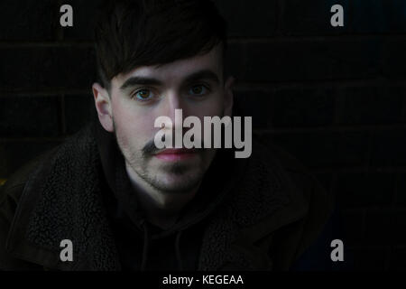 Attractive young adult male against a dark brick wall looking at camera with half of his face shaded Stock Photo