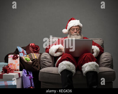 Santa claus relaxing at home and connecting with a laptop, he is chatting and social networking, Christmas time and technology concept Stock Photo