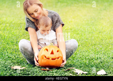 Beautiful young mother with adorable little child, playing with carved pumpkin, enjoying traditional autumn holiday, happy Halloween celebration Stock Photo