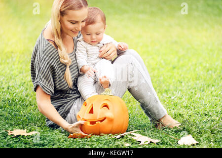 Happy mother and little baby boy outdoors, sitting on green grass field with orange carved pumpkin decorative toy, preparation to Halloween holiday Stock Photo