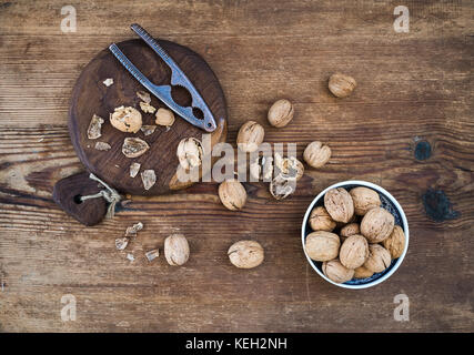 Walnuts in ceramic bowl and on wooden board with nutcracker over rustic wooden background, top view Stock Photo