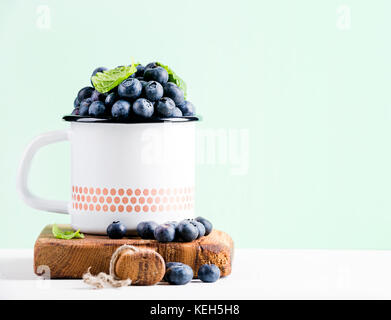 Fresh ripe blueberries in country style enamel mug on rustic wooden board over mint pastel background, selective focus Stock Photo
