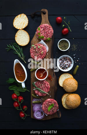Ingredients for cooking burgers. Raw ground beef meat cutlets on wooden chopping board, buns, red onion, cherry tomatoes, greens, pickles, tomato sauc Stock Photo