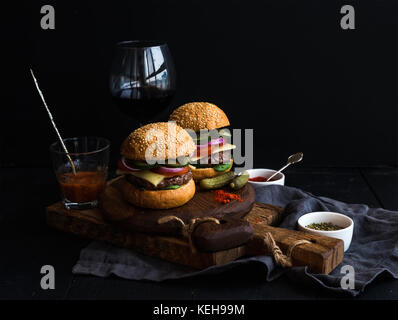 Fresh beef burgers on rustic wooden boards with glass of wine and tomato sauce, black background, selective focus Stock Photo