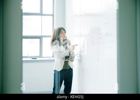 Caucasian businesswoman holding digital tablet and writing on whiteboard Stock Photo