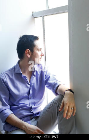 Pensive Caucasian man sitting on windowsill Stock Photo