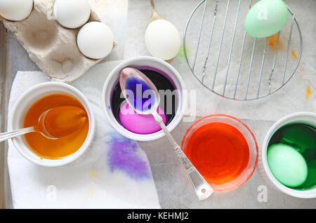 Various bowls with dye, eggs being dyed, spoons, a small rack for drying with dye stains on paper towels underneath Stock Photo
