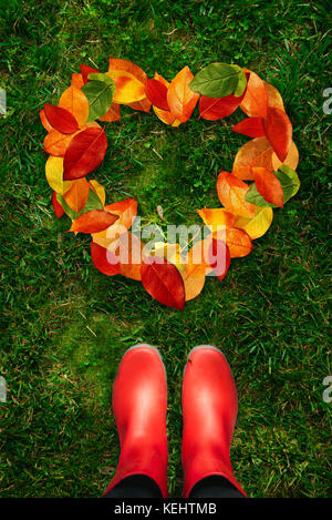 High angle view of a woman in red rubber boots looking at shape of heart created by fallen autumn leaves lying on green grass. Stock Photo
