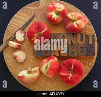 Fresh donut peaches, whole and with pieces cut, with words in wood type and a kitchen knife on a cutting board Stock Photo
