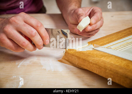 Close-up A male carpenter in work clothes restoring a vintage beige door with wax and a spatula on a wooden table Stock Photo