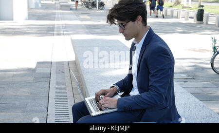 Handsome young man sitting on a stone bench working outdoors in an urban setting typing information onto his laptop computer Stock Photo
