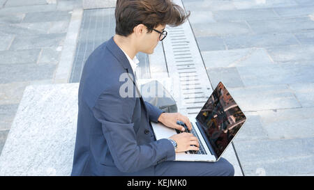Handsome young man sitting on a stone bench working outdoors in an urban setting typing information onto his laptop computer Stock Photo