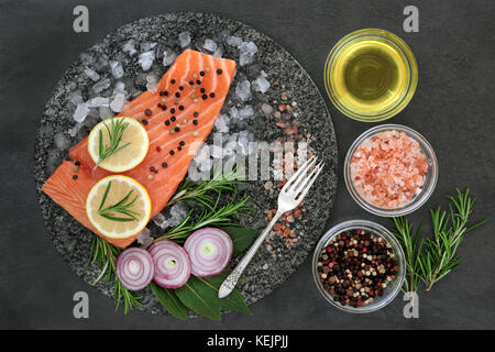 Salmon on crushed ice with herbs, himalayan salt, lemon fruit, onion, peppercorns and olive oil on marble slab on slate background. Healthy heart food. Stock Photo