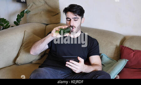 Young attractive man sitting on couch at home in living-room, drinking beer and reading book from ebook reader, relaxing in a lazy afternoon Stock Photo