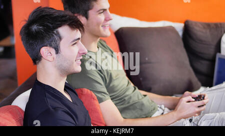 Two young men in casual chilling on sofa and both playing video game console with joysticks in hands Stock Photo