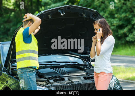 Man Looking At Woman Talking On Mobile Phone With Broken Down Car On Street Stock Photo