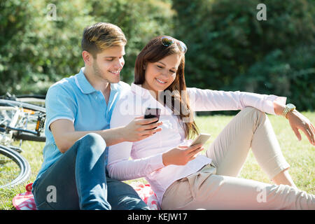 Happy Young Couple Looking At Mobile Phone In Park Stock Photo