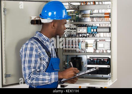 Male Technician Holding Clipboard While Examining Fusebox Stock Photo