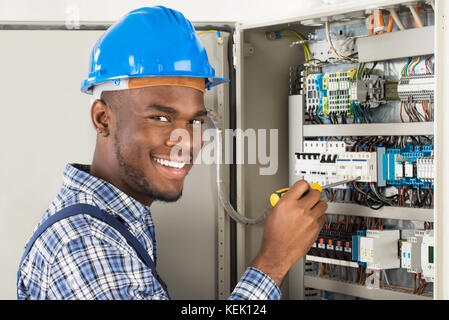 Portrait Of Happy African Male Technician Checking Fusebox With Screwdriver Stock Photo