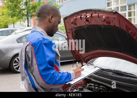 Young African Mechanic Writing On Clipboard In Front Of Open Car Engine Stock Photo