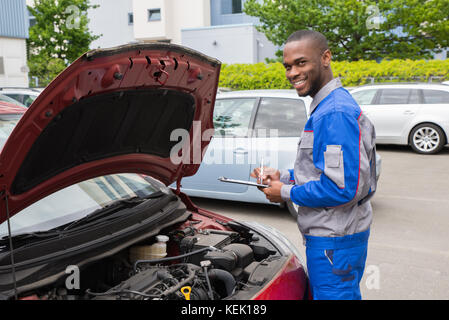 Young African Mechanic Writing On Clipboard In Front Of Open Car Engine Stock Photo