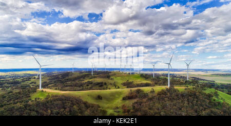 A farm of wind turbine generating renewable electricity high on hill top in rural NSW under blue cloudy sky on a sunny day. Stock Photo