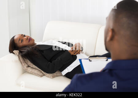 African American Woman Laying On Couch In Front Of Psychiatrist With Clipboard Stock Photo