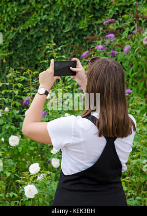 A young girl with a mobile phone photographing a green garden, with a watch on her hands, in a white shirt and a black skirt Stock Photo