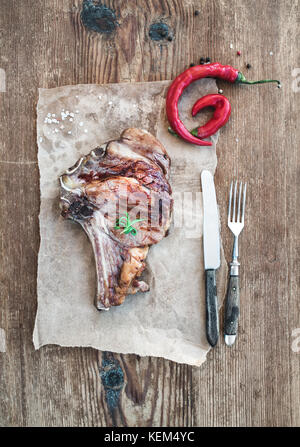 Cooked meat ribeye steak with spices, red chili and rosemary on piece of craft paper over rustic wooden background, top view. Stock Photo