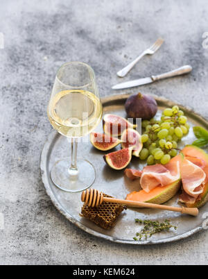 Wine appetizer set. Glass of white wine, honeycomb with drizzlier, figs, grapes, melon and prosciutto on silver tray over rustic grunge surface.  Stock Photo