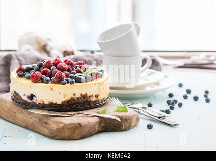 Cheesecake with fresh raspberries and blueberries on a wooden serving board, plates, cups, kitchen napkin, silverware over blue background, window at the backdrop. Stock Photo