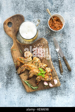 Fried chicken wings on rustic serving board, spicy tomato sauce, herbs and mug of light beer over black wooden backdrop Stock Photo