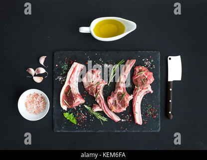 Raw lamb chops. Rack of Lamb with garlic, rosemary and spices on black slate tray, oil in a saucer, salt, dinnerware over dark rustic wood background. Stock Photo
